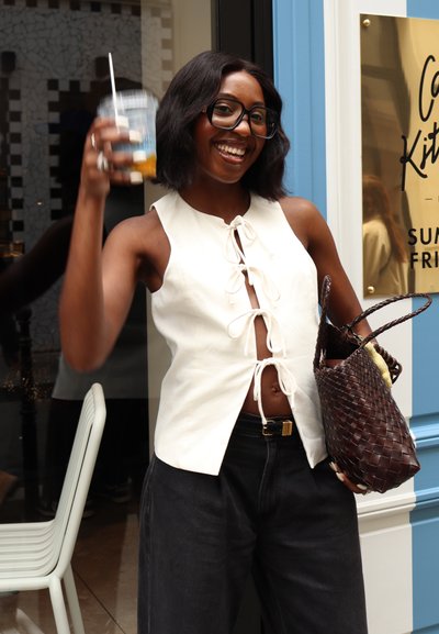 Una blusa sin mangas blanca con un detalle de lazo en la parte delantera, combinada con unos pantalones vaqueros de pierna ancha y tiro alto negros. Un bolso de mano tejido marrón y una bebida fría.