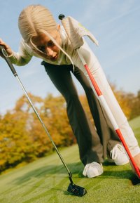 Schwarzer Golf-Putter mit roten Akzenten auf grünem Gras. Der Golfer trägt graue Hosen und ein eng anliegendes weißes Langarmshirt und beugt sich über den Putter.