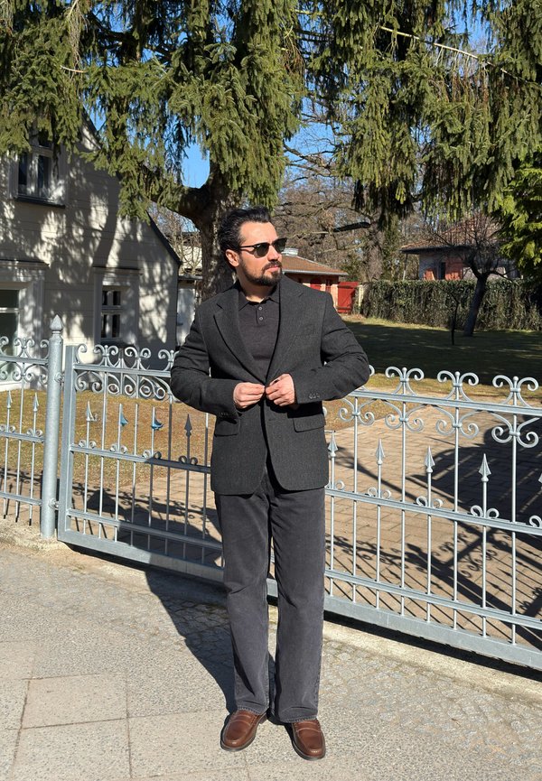 Homme portant un blazer sombre, un pantalon noir et des lunettes de soleil, debout devant une grille en métal avec des arbres et des maisons en arrière-plan.