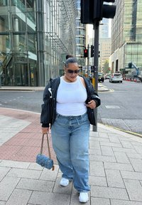 Visual descontraído com uma t-shirt branca sem mangas, uma jaqueta oversized às riscas pretas, jeans baggy azul claro e ténis brancos, complementado com óculos de sol e um colar de pérolas.