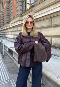 Brązowa, skórzana, oversize'owa kurtka, ciemny, dziergany top i ciemne spodnie dżinsowe. Trzyma brązową zamszową torbę ze ściągaczem. Miejski krajobraz w tle.