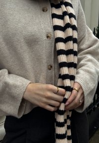 Beige Strickjacke mit großen Knöpfen, kombiniert mit einem schwarz-creamfarbenen gestreiften Schal. Die Accessoires umfassen mehrere Ringe an den Fingern.