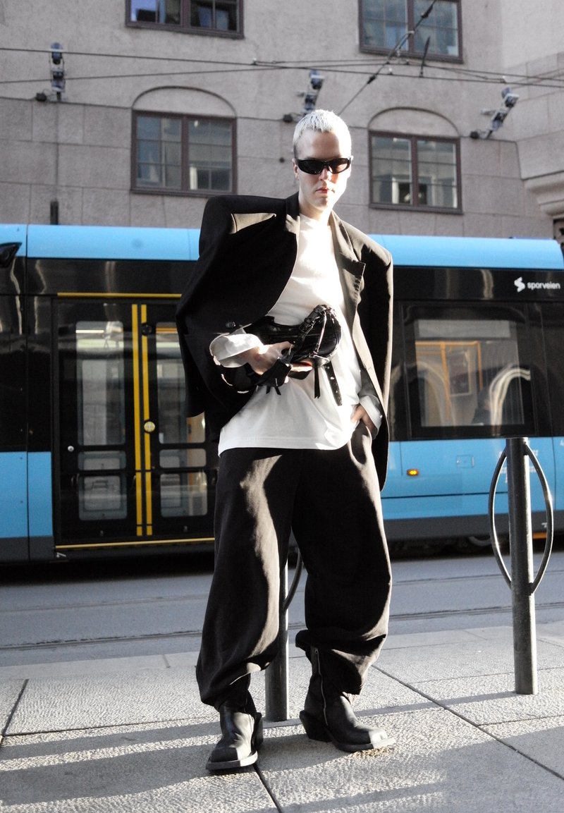 Personne aux cheveux blonds courts portant des lunettes de soleil noires, un blazer noir oversize, une chemise blanche et un pantalon noir, debout devant un tram bleu.