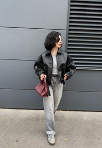 Donna con capelli ricci che indossa una giacca di pelle nera, jeans grigi e sneaker grigie, mentre tiene una borsa bordeaux, in piedi di fronte a un muro grigio.