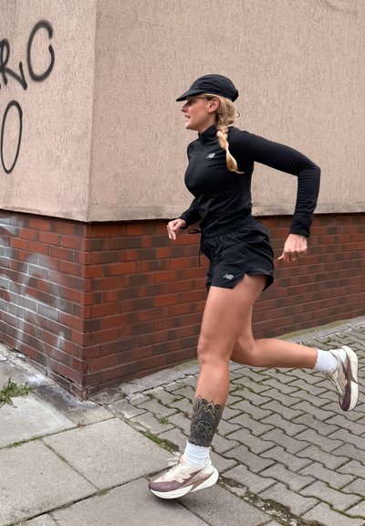Camiseta negra de manga larga, pantalones cortos negros y zapatillas de correr gris-marrón con calcetines blancos. Tatuaje visible en la pantorrilla con detalles. Postura de carrera.