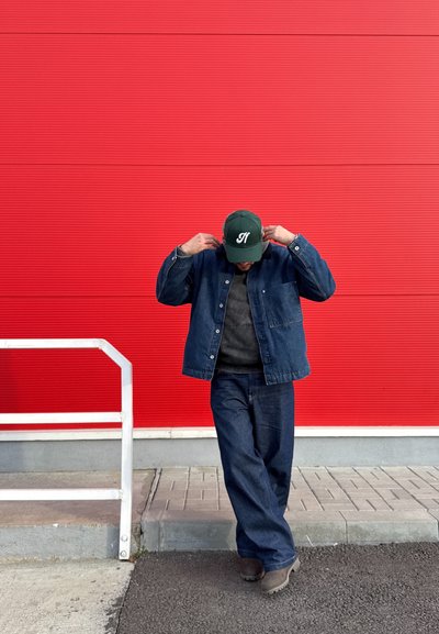 Chaqueta y pantalones de mezclilla en azul oscuro, suéter gris debajo, gorra verde con logo blanco, de pie frente a una pared roja con textura.