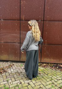 Pull gris en maille torsadée à manches longues, associé à un pantalon noir à larges jambes à fines rayures. Fond texturé avec des panneaux couleur rouille.