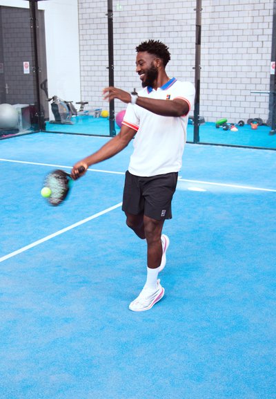 Jugador de pádel con una polo blanca con acentos rojos, pantalones cortos negros y zapatillas deportivas, agitando una raqueta en una pista azul con texturas claras.