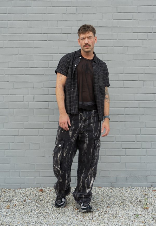 Homme avec moustache et tatouages debout contre un mur en briques grises, portant une chemise noire, un pantalon cargo à motifs et des baskets sur un sol gravillonné.