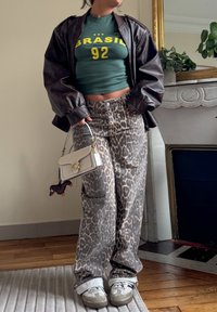 Green cropped t-shirt featuring "BRASIL 92" in yellow, brown leather bomber jacket, grey leopard-print trousers, and grey-white sneakers. Small handbag.