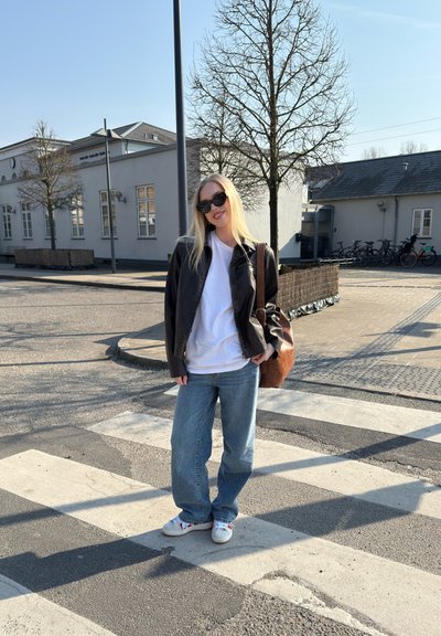 Chaqueta de cuero marrón, camiseta blanca, jeans de mezclilla azules, zapatos blancos con detalles rojos y bolsa de hombro marrón. De pie en un paso de peatones.