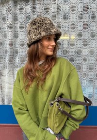 Sweatshirt oversized verde, chapéu de pele com padrão de leopardo, bolsa verde-oliva com uma alça de couro. O fundo apresenta uma cortina de renda estampada.