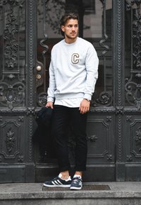 Szürke pulóver "C" logóval, fekete nadrág és szürke Adidas sportcipő. Díszes fémajtó előtt, bonyolult mintázattal.