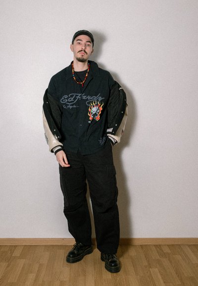 Chaqueta de estilo universitario negra con gráficos de llamas y calaveras, mangas beige, combinada con pantalones cargo negros y zapatos negros. Accesorio de collar de cuentas.