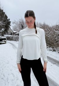 Haut de sport blanc à manches longues avec encolure ronde, tissu léger et coupe décontractée, associé à un legging noir dans un environnement enneigé.
