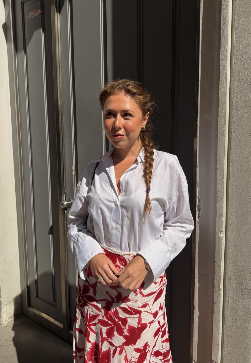 Jeune femme aux cheveux tressés, portant un chemisier blanc et une jupe rouge fleurie, debout au soleil près d'une porte grise.