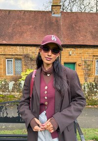 Femme portant une casquette LA bordeaux, des lunettes de soleil assorties, des vêtements superposés marron et bordeaux, et un pantalon gris, debout devant une maison en pierre avec un toit rouge.