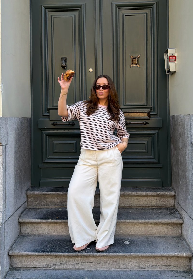 Femme en chemise rayée et pantalon blanc debout sur des marches en pierre, tenant un croissant en l'air, devant une grande porte vert foncé.