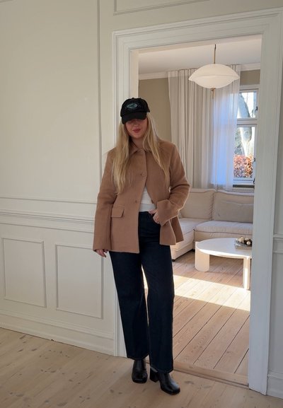 Abrigo de lana marrón con cuello, llevado sobre una blusa blanca; combinado con jeans de mezclilla oscura y botas negras de tobillo. Gorra negra, piel clara.