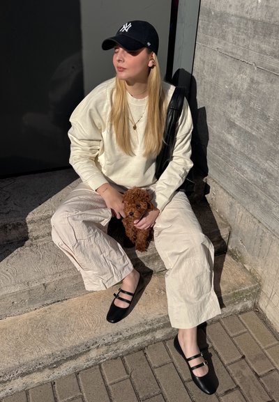 Sudadera crema de gran tamaño, pantalones anchos beige texturizados, zapatos planos negros de doble tira y un pequeño perro marrón en los escalones bajo la luz del sol.