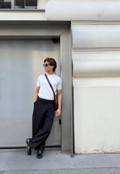 Camiseta blanca de manga corta, pantalones holgados negros con un botón y zapatos negros. Los accesorios incluyen gafas de sol y un bolso de hombro marrón.