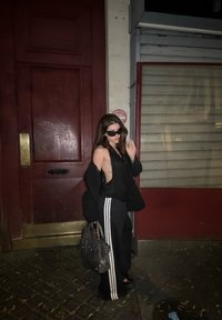 Conjunto preto composto por uma blusa sem mangas e calças de perna larga com riscas brancas. A figura leva uma mala cinza e usa óculos de sol escuros.