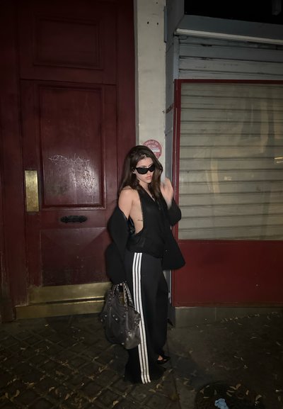 Conjunto negro que presenta una blusa sin mangas y pantalones de pierna ancha con rayas blancas. La figura lleva un bolso gris y usa gafas de sol oscuras.