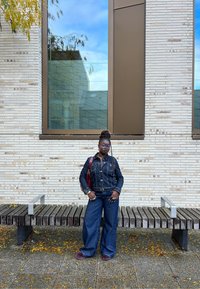 Jeans jakna in široke hlače, obe v temno modri barvi. Dodatki vključujejo burgundsko torbo. Ozadje prikazuje opečno steno in veliko okno.
