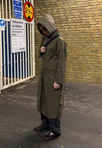 Palton lung verde măsliniu cu glugă, pantaloni închiși la culoare și pantofi maro. Persoana stă lângă un zid de cărămidă, cu semne de parcare vizibile.