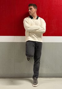 Krém pulóver sötét gallérral, szürke farmernadrág és szürke cipők. Egy piros-szürke falnak támaszkodva, karját keresztbe téve, oldalra néz.