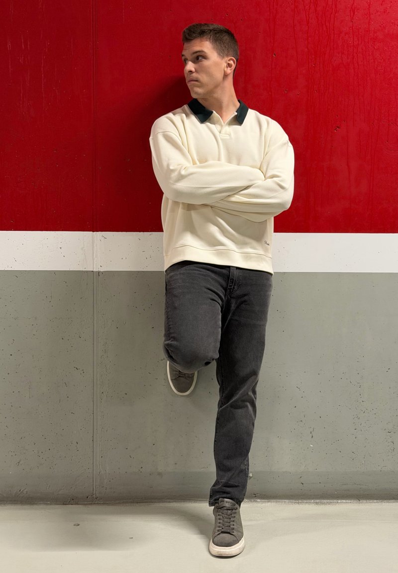 Sweat-shirt crème avec col sombre, jeans gris et chaussures grises. Appuyé contre un mur rouge et gris, les bras croisés et regardant de côté.