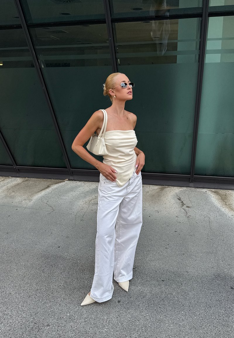 Femme aux cheveux blonds en chignon, portant des lunettes de soleil, un top crème sans bretelles, un pantalon blanc à jambes larges et des chaussures blanches pointues, debout à l'extérieur.