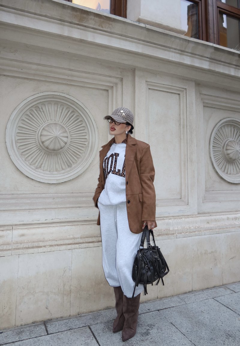 Femme en blazer marron, pantalon de survêtement blanc, casquette à carreaux et bottines marron, tenant un sac à main noir, debout contre un mur en pierre ornée.