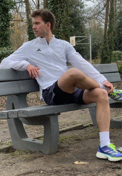 Hombre atlético sentado en un banco de concreto, vistiendo una chaqueta blanca con cremallera, pantalones cortos negros y zapatillas multicolores con acentos tonales.