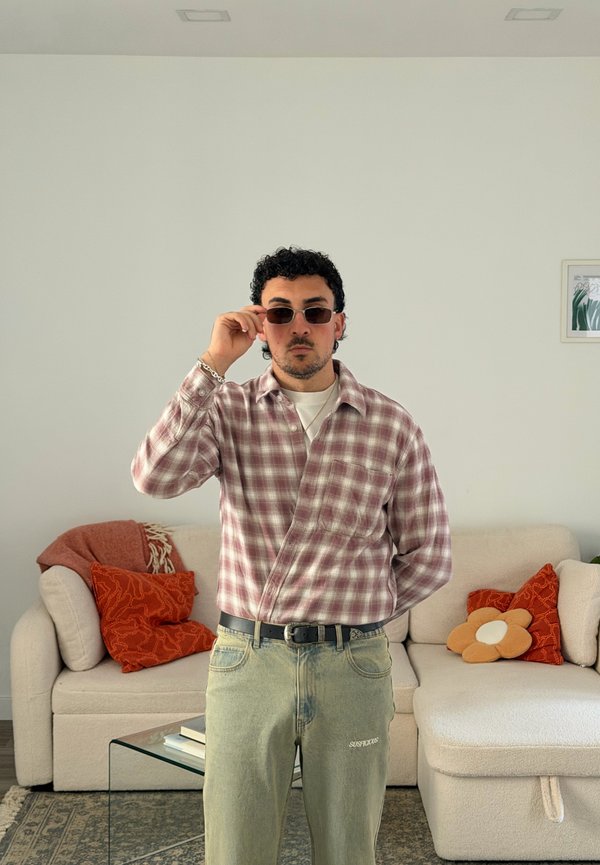 Mann mit Sonnenbrille, kariertem Hemd und hellen Jeans steht in einem Wohnzimmer mit einem beigen Sofa und bunten Kissen.