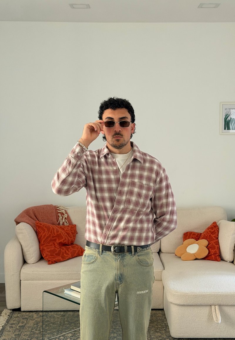 Homem a usar óculos de sol, camisa xadrez e jeans claros, em pé numa sala com um sofá bege e almofadas coloridas.