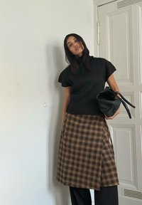 Fekete bordázott felső rövid ujjal, barna kockás átlapolt szoknyával kombinálva. Egy fekete táskát tart, semleges háttér előtt.