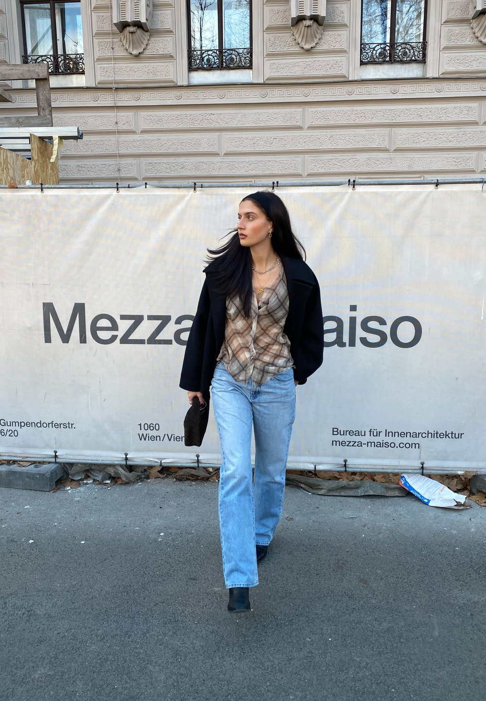 Modelo com um casaco preto, camisa xadrez e jeans azul-claro, segurando uma mala preta, a andar numa rua da cidade ao lado de uma barreira de construção.