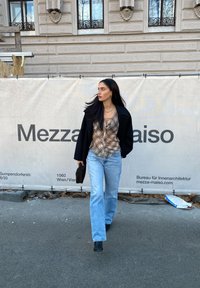 Modelo com um casaco preto, camisa xadrez e jeans azul-claro, segurando uma mala preta, a andar numa rua da cidade ao lado de uma barreira de construção.