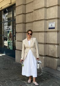 Μπεζ σακάκι με καλή εφαρμογή πάνω από ένα άσπρο maxi φόρεμα, κρατώντας μια διάφανη τσάντα και ένα ποτό. Τα γυαλιά ηλίου και το ελαφρύ υπόδ ημα συμπληρώνουν την εμφάνιση.