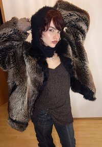 Manteau en fausse fourrure avec une capuche noire, présentant un mélange de textures grises et brunes. Porté sur un haut foncé à motifs et un pantalon gris.