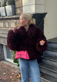 Mulher com cabelo loiro em rabo de cavalo, vestindo uma jaqueta felpuda bordô e jeans azuis, segurando um bálsamo labial perto dos lábios, parada ao lado de escadas ao ar livre.