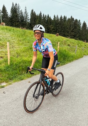 Ciclista su una bici da corsa nera con finitura opaca, che indossa una maglietta geometrica multicolore e pantaloni neri, pedala su un sentiero asfaltato.