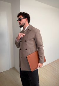 Barna gyapjú kétsoros kabát sötét gombokkal, fekete nadrággal párosítva. Egy sima, barna bőrből készült laptoptokot tart egy semleges környezetben.