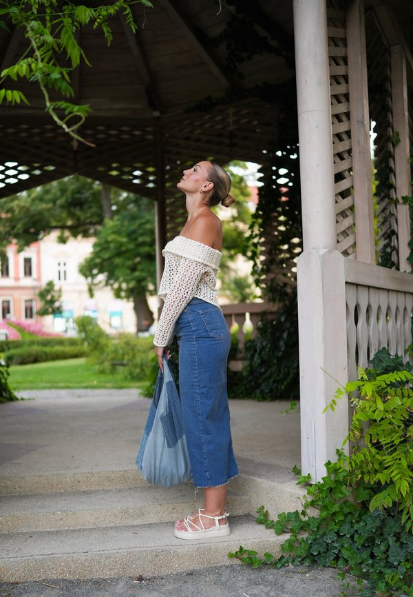 Vállnélküli krémszínű kötött blúz, magas derekú kék denim szoknya, bézs pántos szandál, kezében egy textúrált kék táska, lépcsőkön áll, zöld növényzet mellett.