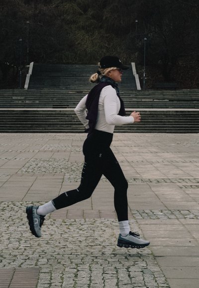 Atleta con camiseta blanca de manga larga y mallas negras, llevando gorra y zapatillas de correr, trotando por un camino de piedra texturizada.
