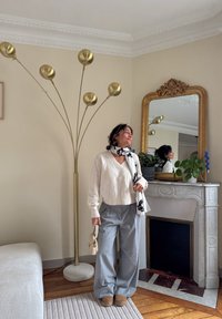 Pull en maille blanc, pantalon gris à jambes larges, écharpe noire et blanche. Lampe sur pied dorée, miroir orné, cheminée en marbre, plante en pot. Sol en bois.