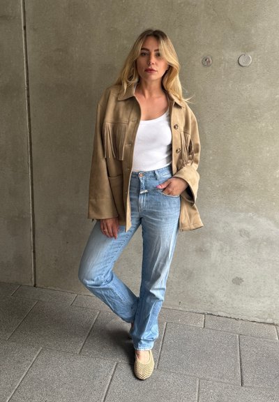 Chaqueta de ante marrón con detalles de flecos, top ajustado blanco, jeans de mezclilla azul claro y zapatos de yute beige. Fondo sencillo.