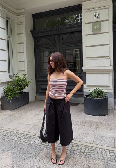 Mujer con top tipo tubo a rayas, pantalones negros de pierna ancha, chanclas y gafas de sol, sosteniendo un bolso negro, de pie junto a una puerta negra con plantas cerca.