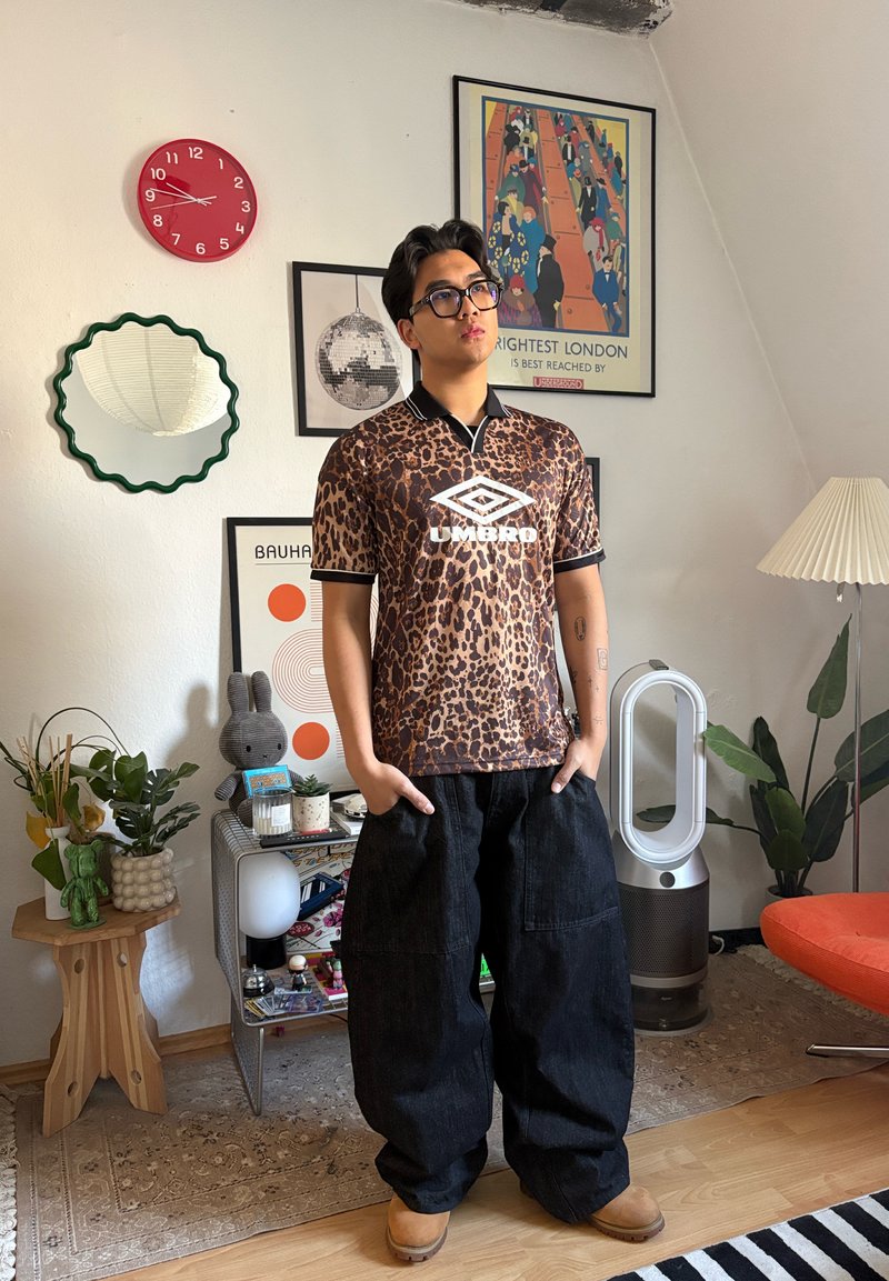 Jeune homme portant des lunettes, un maillot Umbro à motif léopard et un pantalon large noir, debout dans une pièce lumineuse décorée de plantes, d'un miroir, de posters et d'une horloge.